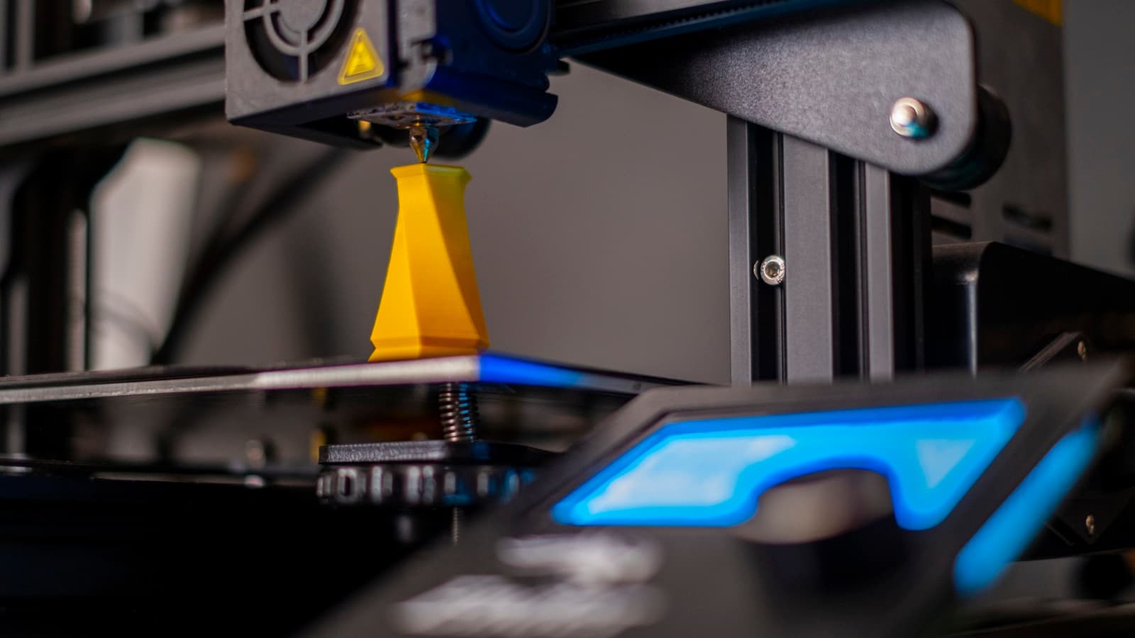 Close-up of a 3D printer extruder finishing a yellow printed part with a blue-lit control screen.