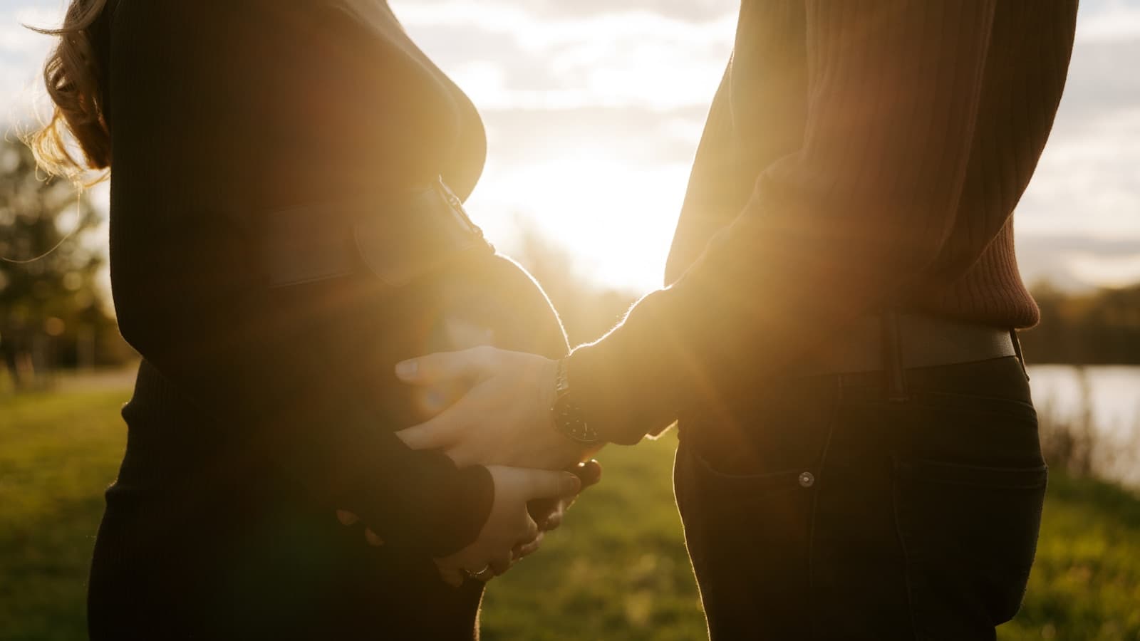 A silhouetted pregnant couple holding hands at golden-hour sunset outdoors.
