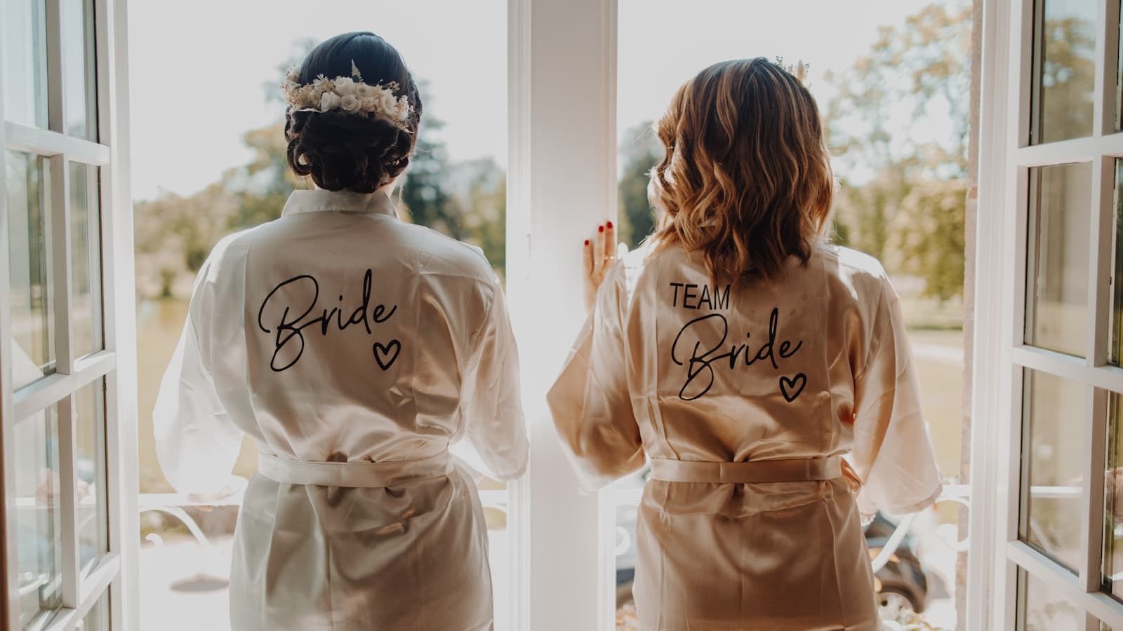 Two bridesmaids in robes looking out a window together.