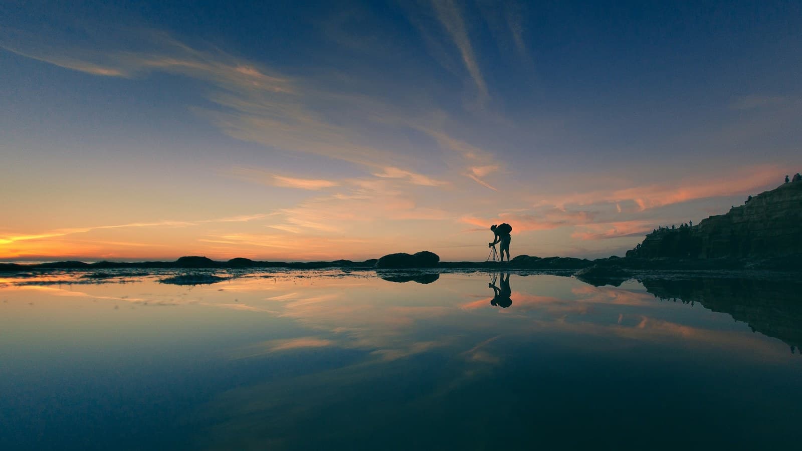 The silhouette of a photographer adjusting a camera on a tripod at sunset over coastal cliffs.