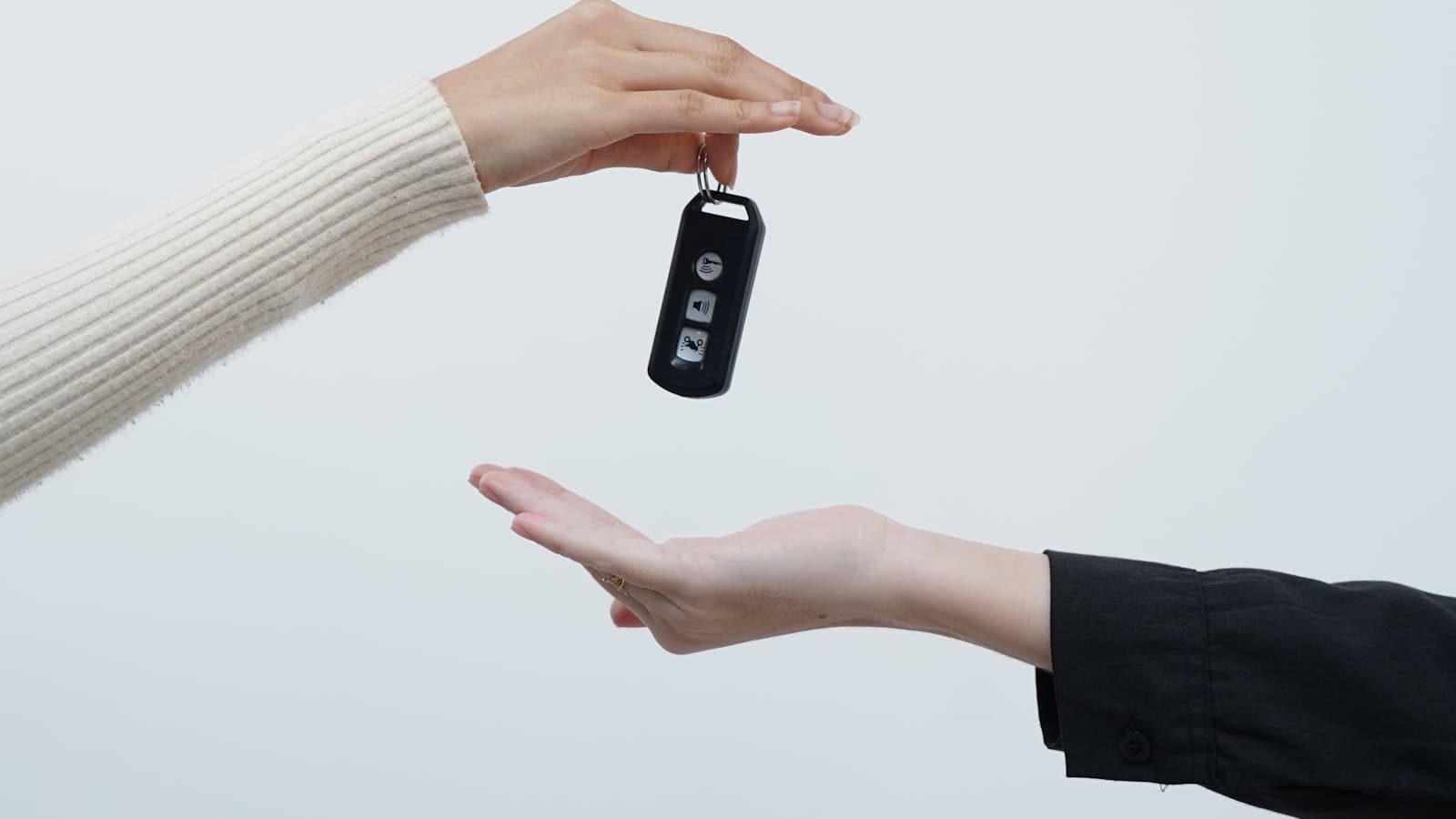 Close-up of two hands exchanging a car key fob against a plain white background, no faces, plates, or legible brand text/logos visible.