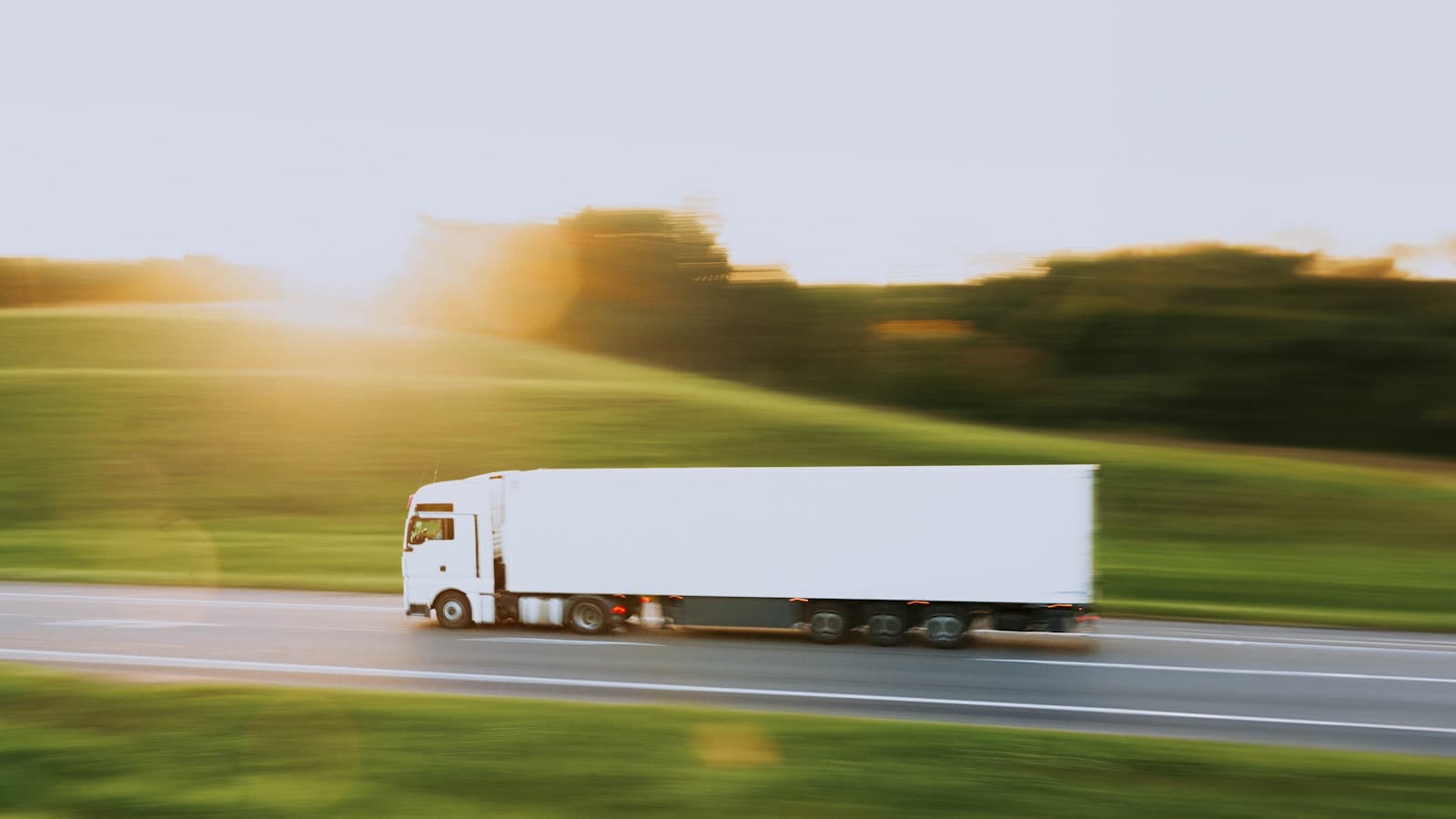 A motion-blurred plain white semi truck speeding down a rural highway at golden hour.