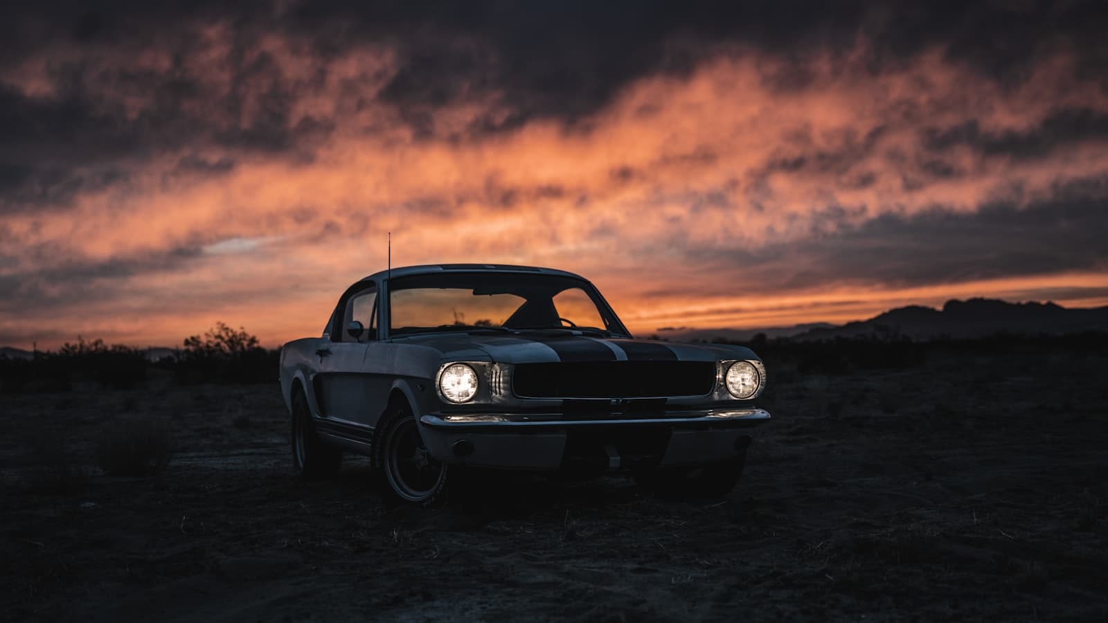 A silhouetted classic muscle car with a racing stripe parked in a desert at sunset with its headlights on against an orange and purple sky.