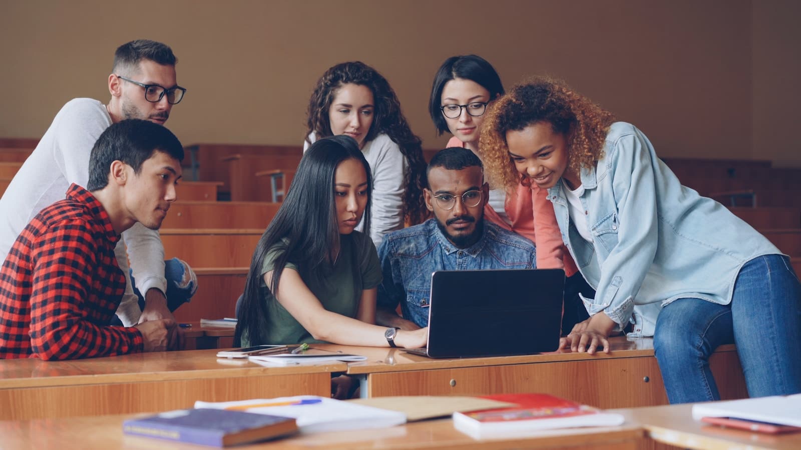 Seven students crowded around a laptop in a classroom, collaborative energy.