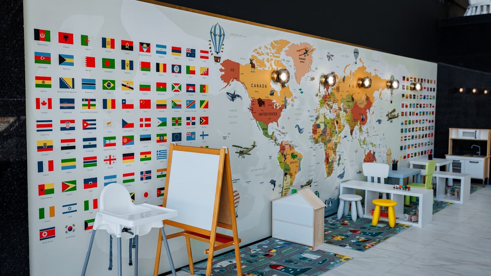 A colorful daycare classroom with small tables, chairs, storage cubes, and a world-map wall mural.