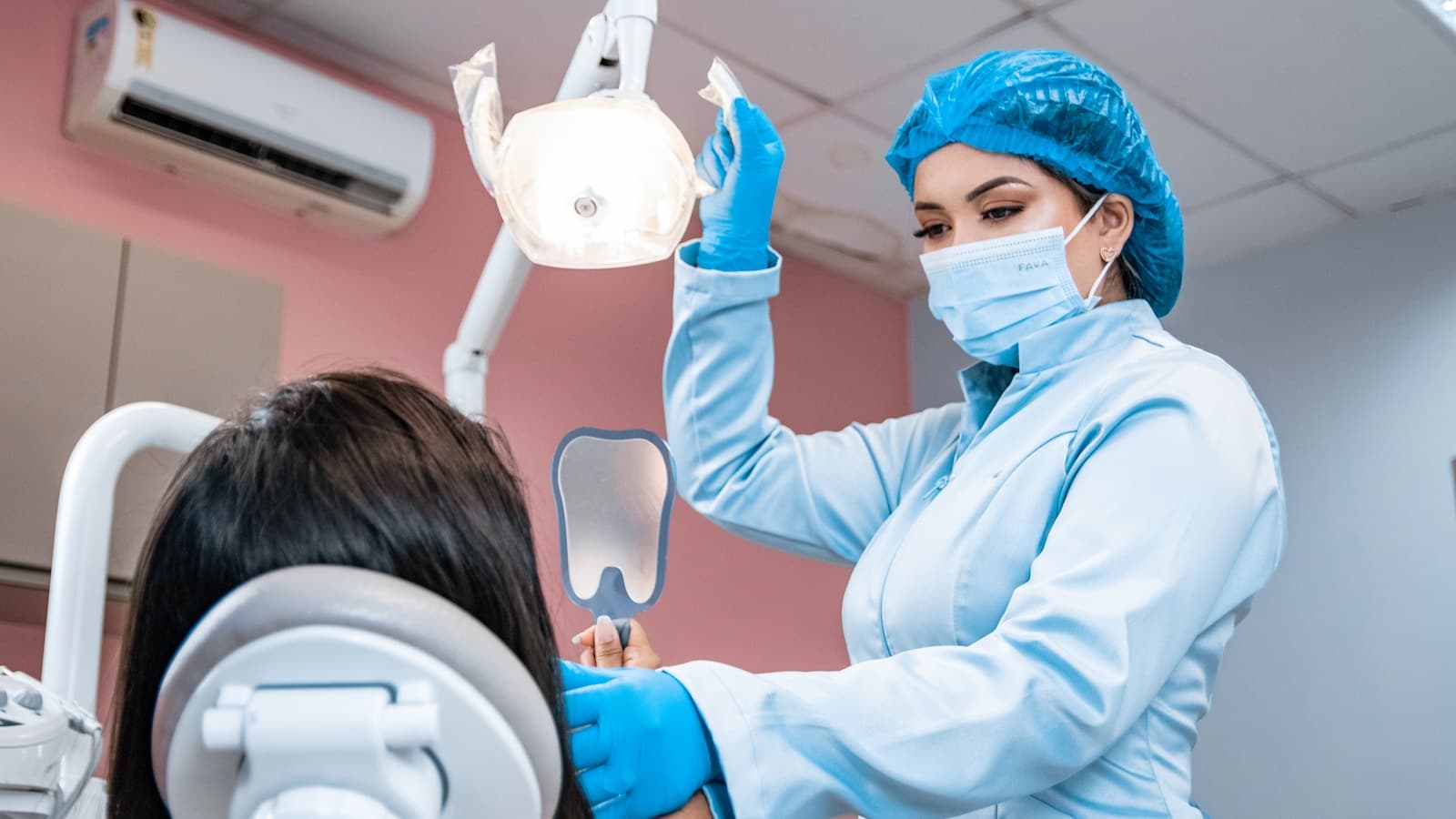 A dental hygienist in blue scrubs adjusting the overhead exam light.