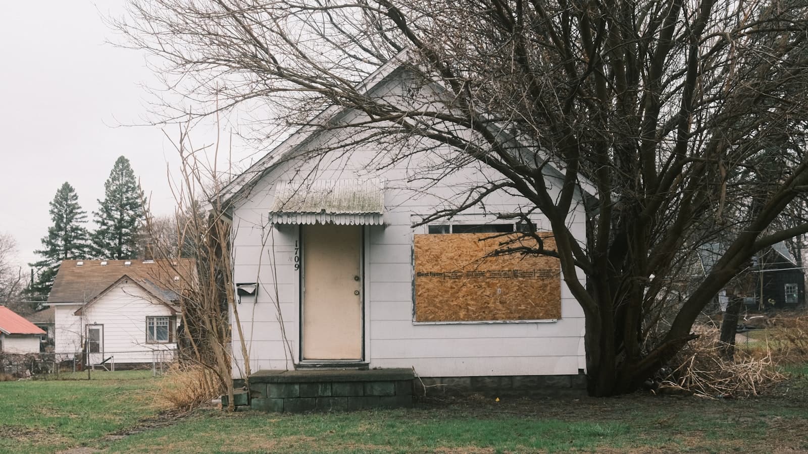 A worn white house with a boarded-up window and bare trees in front, conveying a distressed/foreclosed property with no people or legible signage.