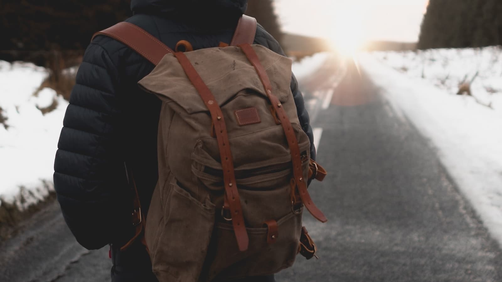 A person seen from behind walking down a snowy road with a backpack toward a sunrise; face not visible.