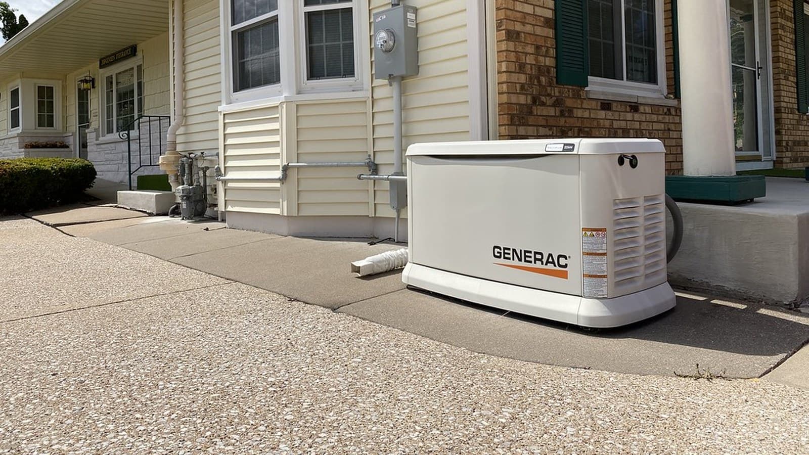 A white Generac standby home generator installed on a pad beside a brick and vinyl-sided house, next to a gas meter and covered porch.