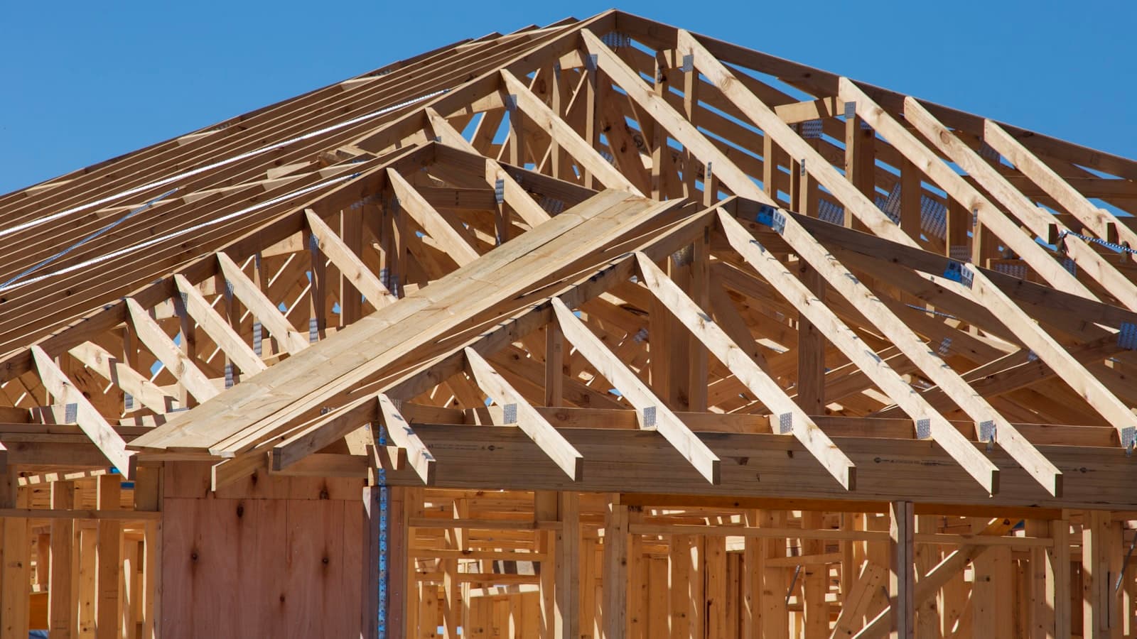 Exposed wooden roof trusses of a house under construction against a clear blue sky, representing a home addition build; no faces or text.