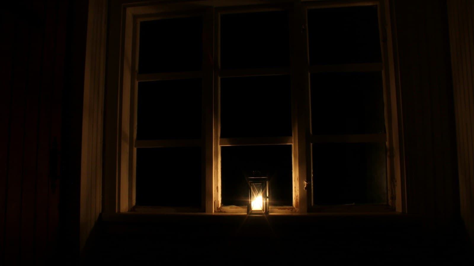 A single lit candle glowing on a windowsill at night, evoking a power outage at home.