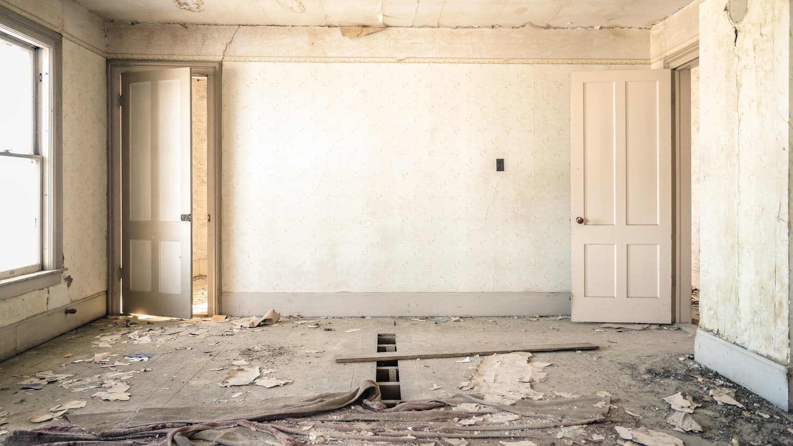 An interior room mid-renovation with peeling wallpaper, a stripped subfloor, and debris scattered about, no people or readable text.