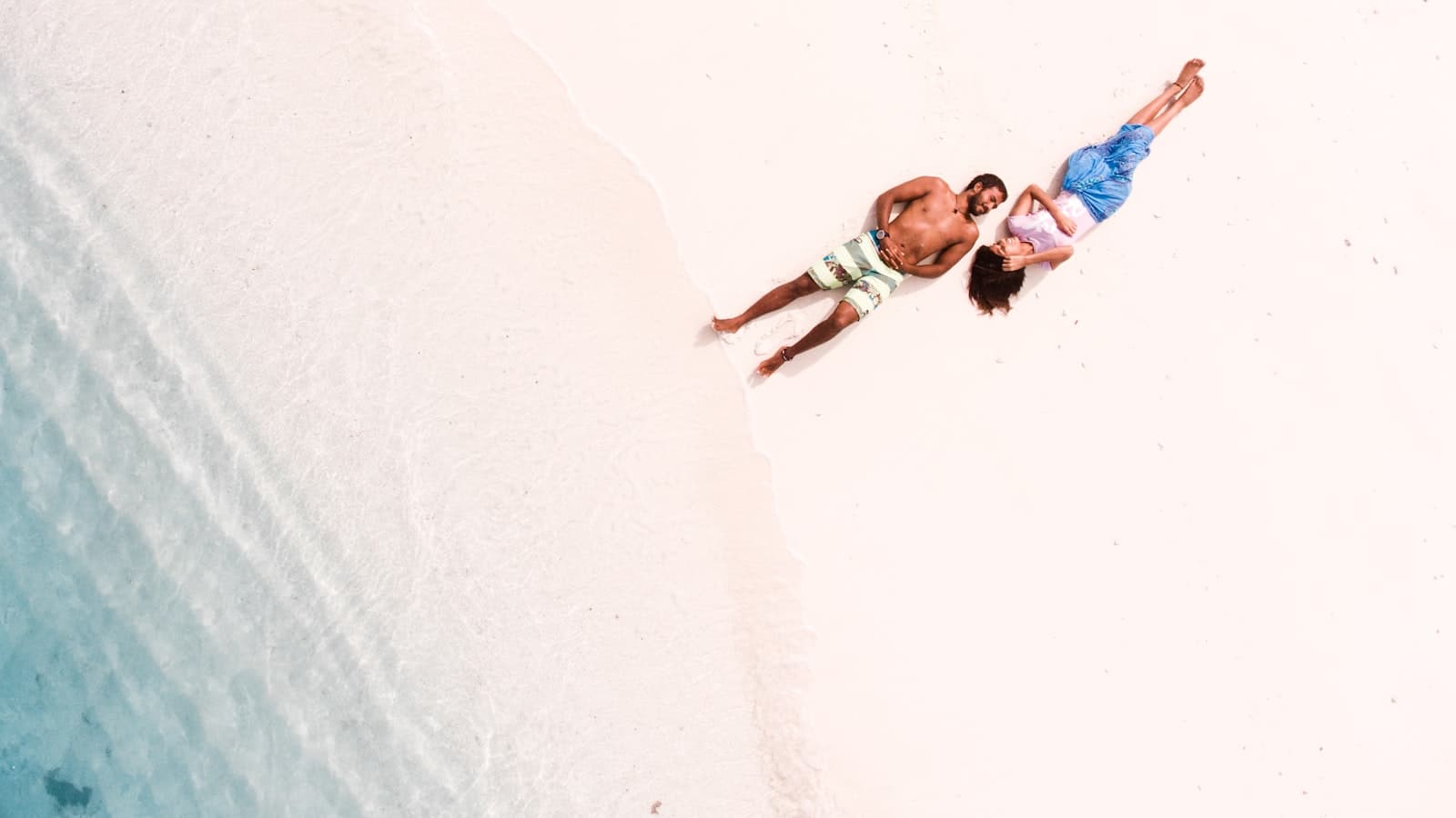 Bird's-eye view of a couple lying together on a white sand beach next to turquoise water, faces not identifiable from directly overhead, no text or logos.