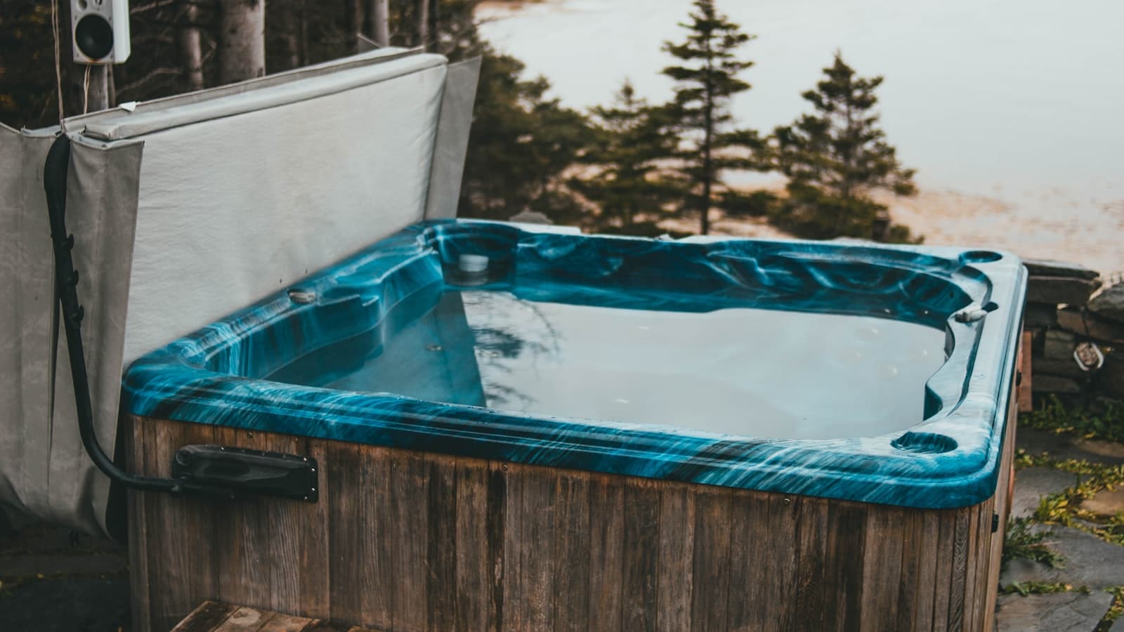 An empty outdoor hot tub with its cover open, surrounded by trees, with no people or readable text visible.
