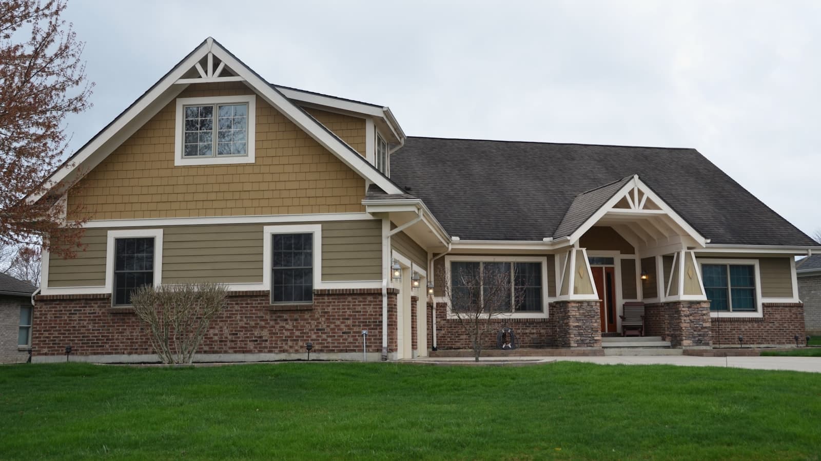 A two-story traditional craftsman-style house with a brick lower level and shake-style gable siding, a covered front porch with stone-based columns, and a fresh green lawn.