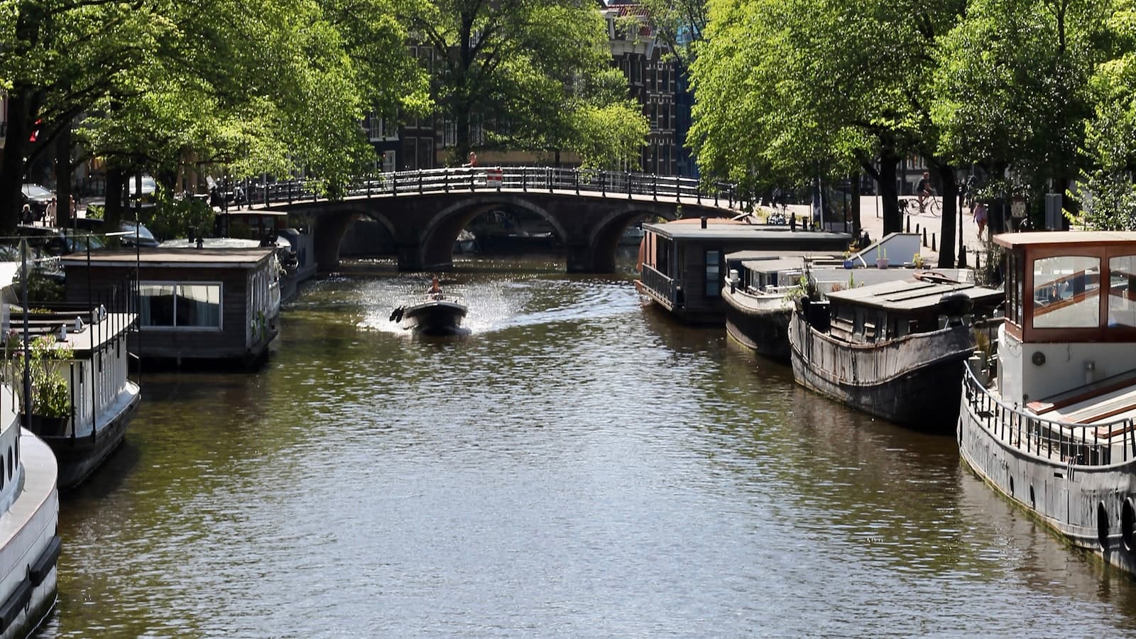 Wooden houseboats moored along a canal beneath a stone bridge with green trees overhead, only small distant unidentifiable figures, no readable text.