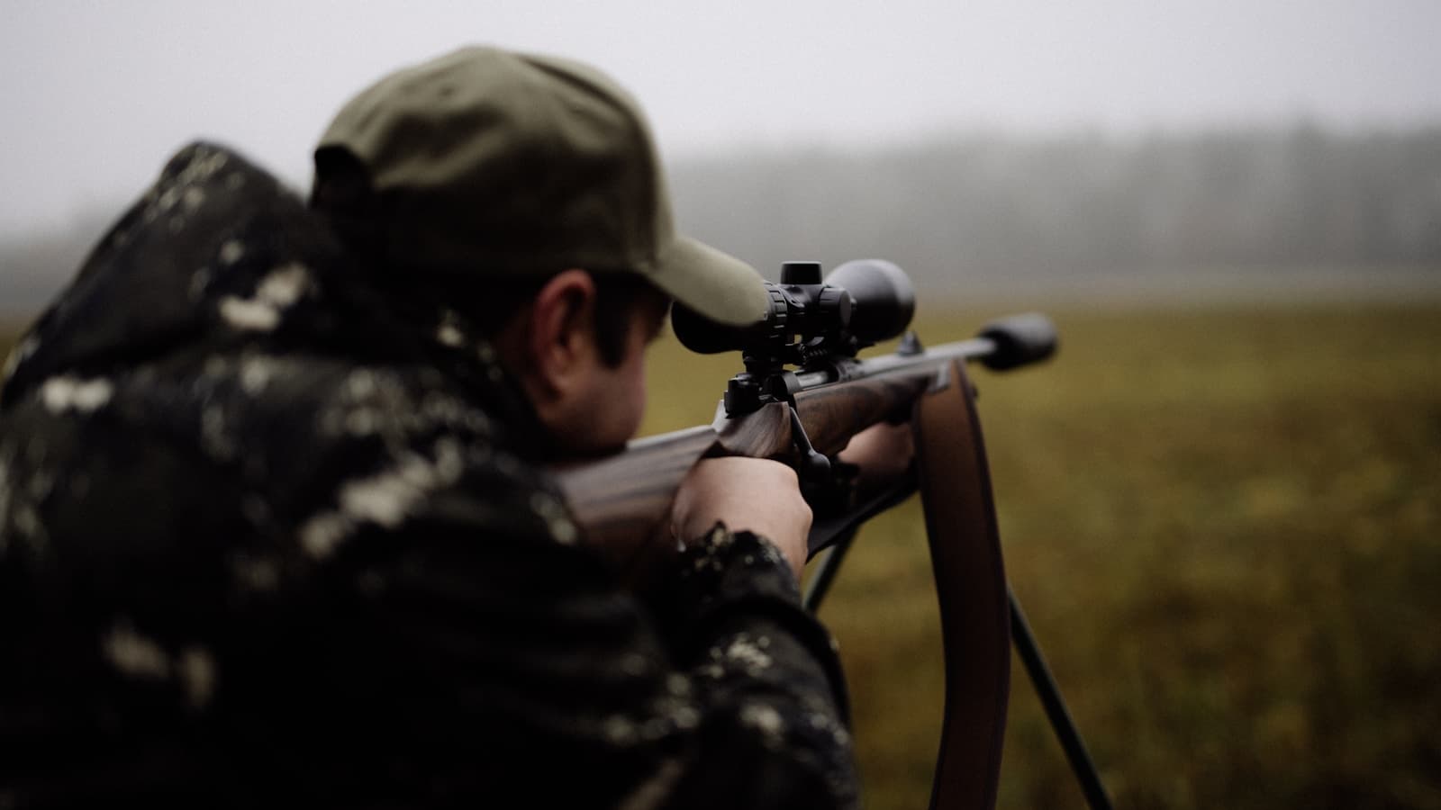 A hunter in a camo and olive cap aiming a rifle across a foggy field.