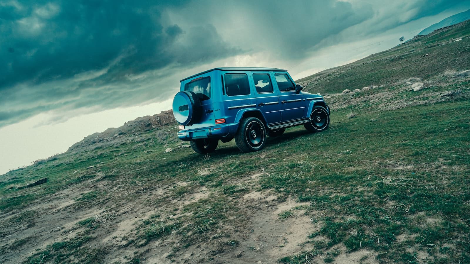 A blue Mercedes G-Class SUV parked on a grassy hillside under a stormy sky, shot from a distance with no readable plate or logo.