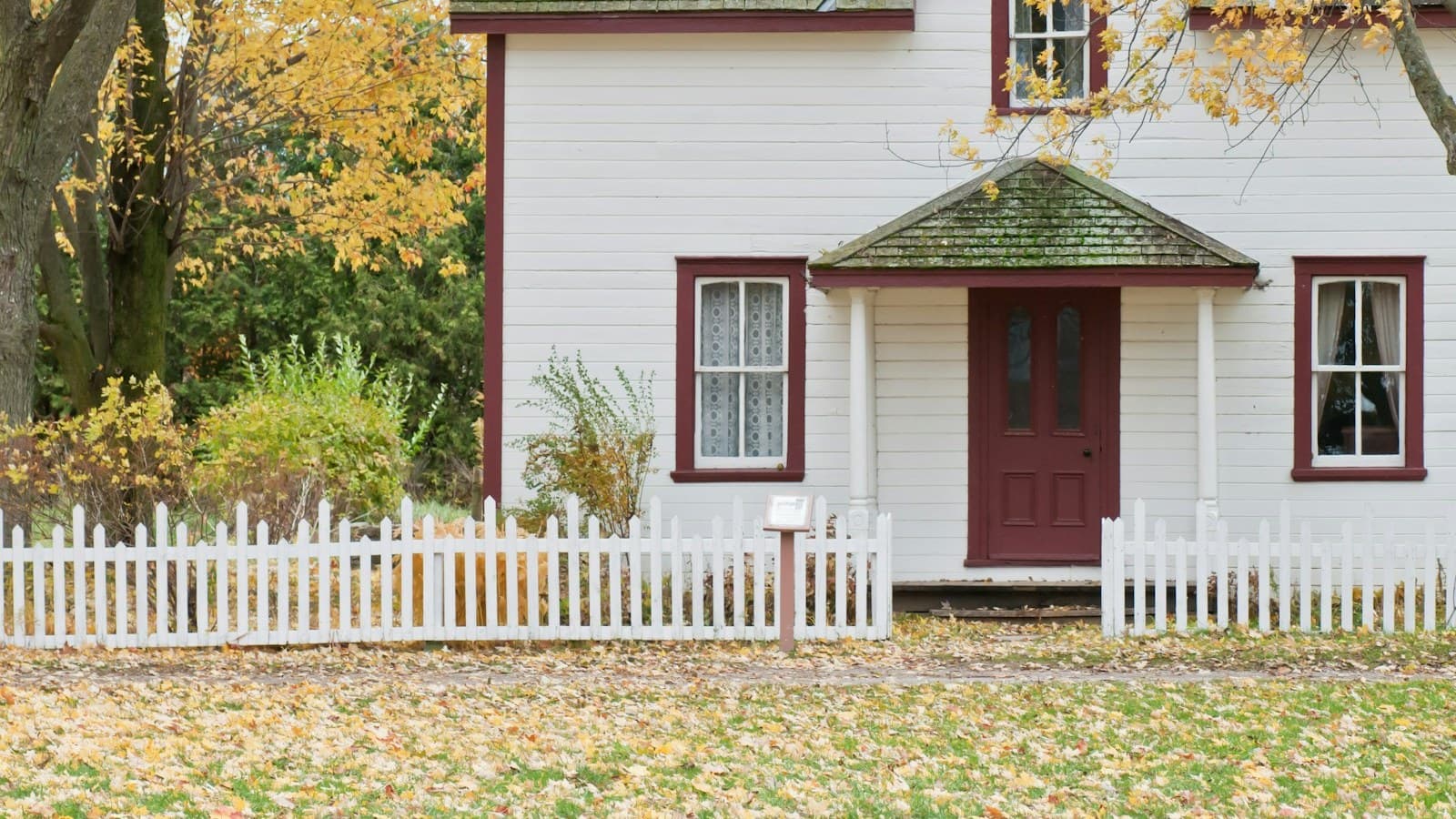 A white house with maroon trim, a white picket fence, and autumn leaves on the lawn, with no people, text, or logos visible.