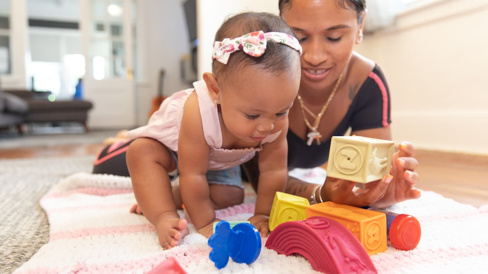 A smiling woman lies on a living room rug helping a baby girl play with colorful stacking toy blocks.