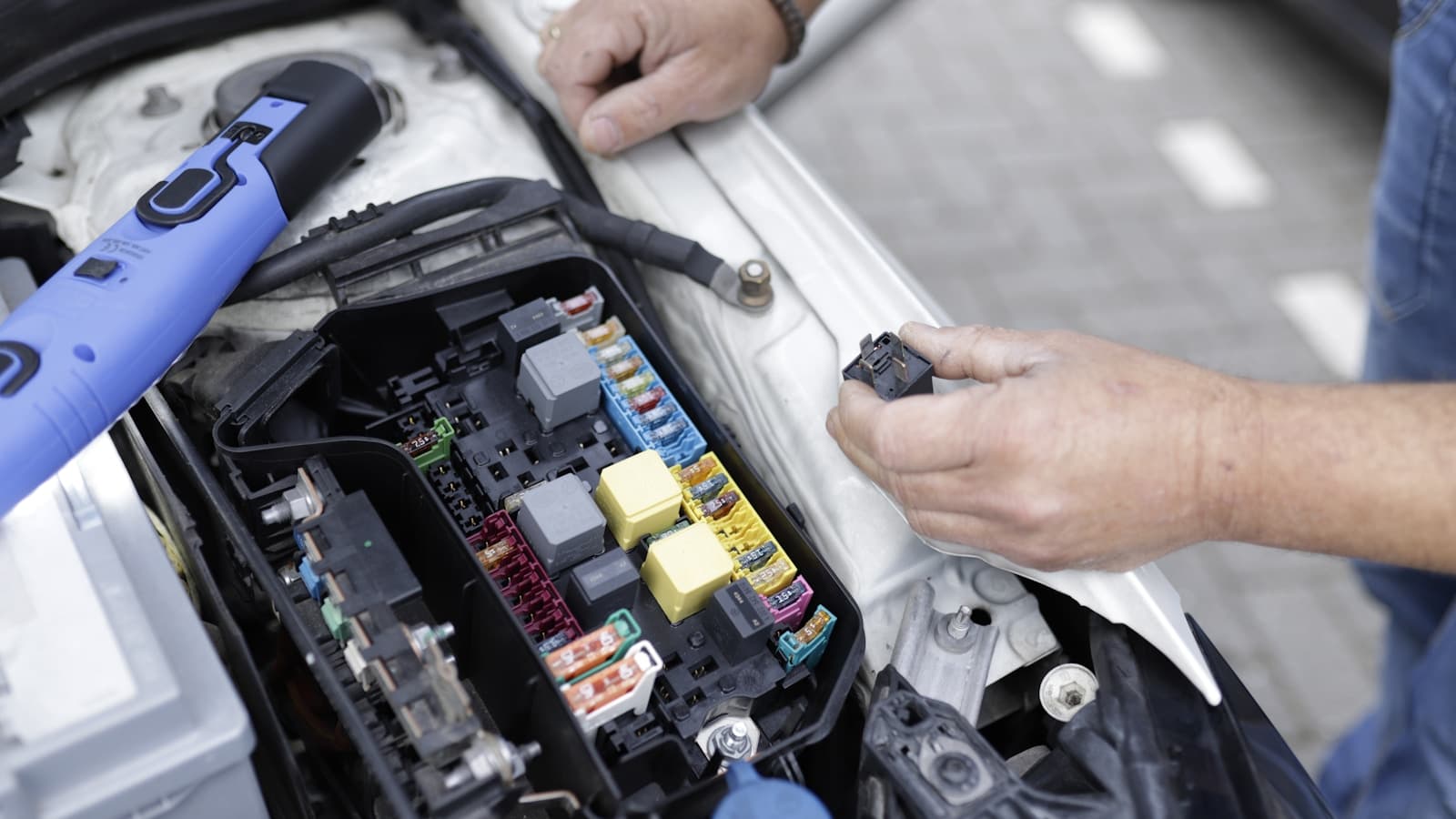 A mechanic working on a car's fuse box and electrical bay under the hood.