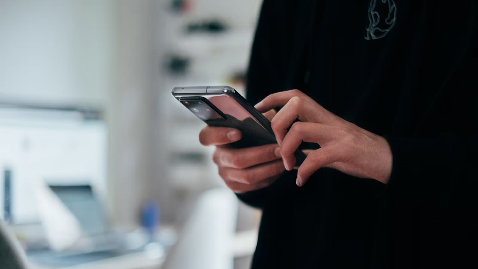 A person's hand holds a modern foldable smartphone indoors, with a blurred desk and laptop visible in the background.