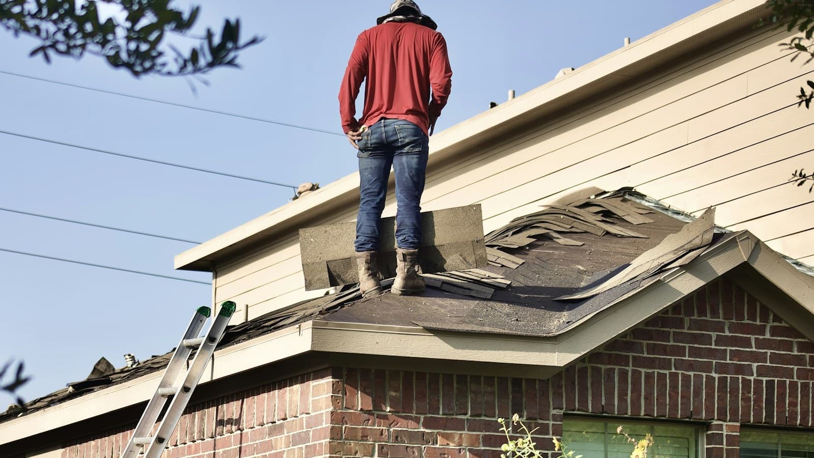 A person (cropped above the shoulders, no face visible) tearing off old shingles on a brick house roof with a ladder propped against the eaves.