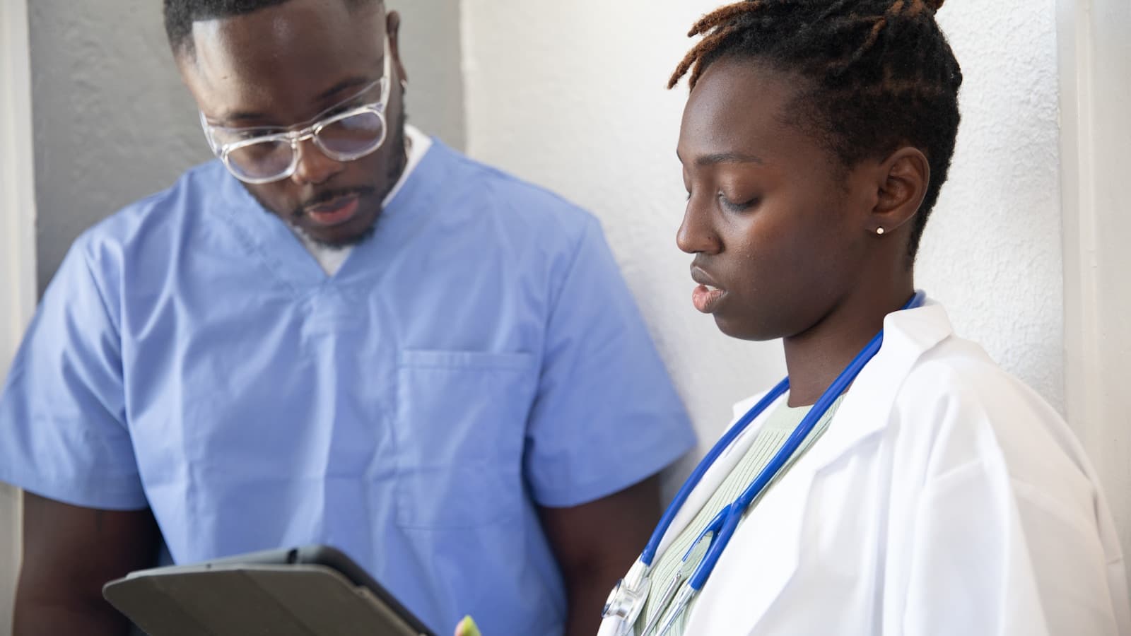 A nurse and doctor reviewing patient info together on a tablet in a clinical hallway.