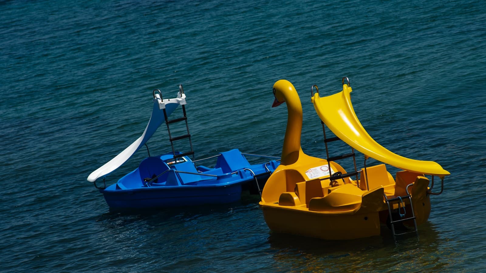 A blue pedal boat and a yellow swan-shaped pedal boat floating side by side on clear blue water.