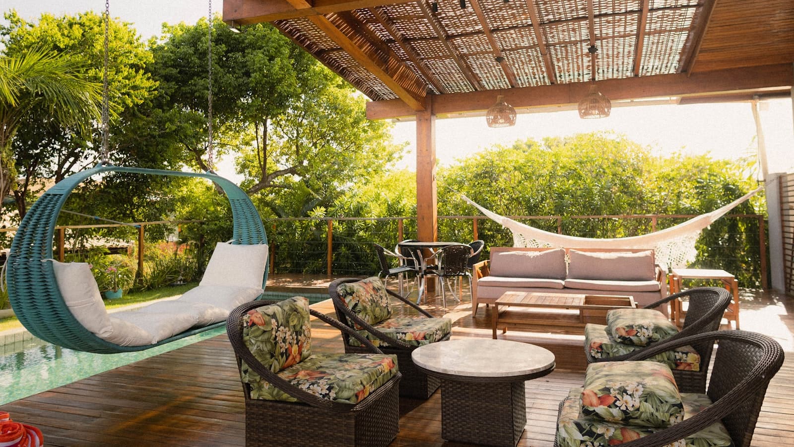 A wood-slatted pergola with a woven roof shades a poolside lounge area with a hanging egg chair, rattan armchairs, a sofa, and a hammock.