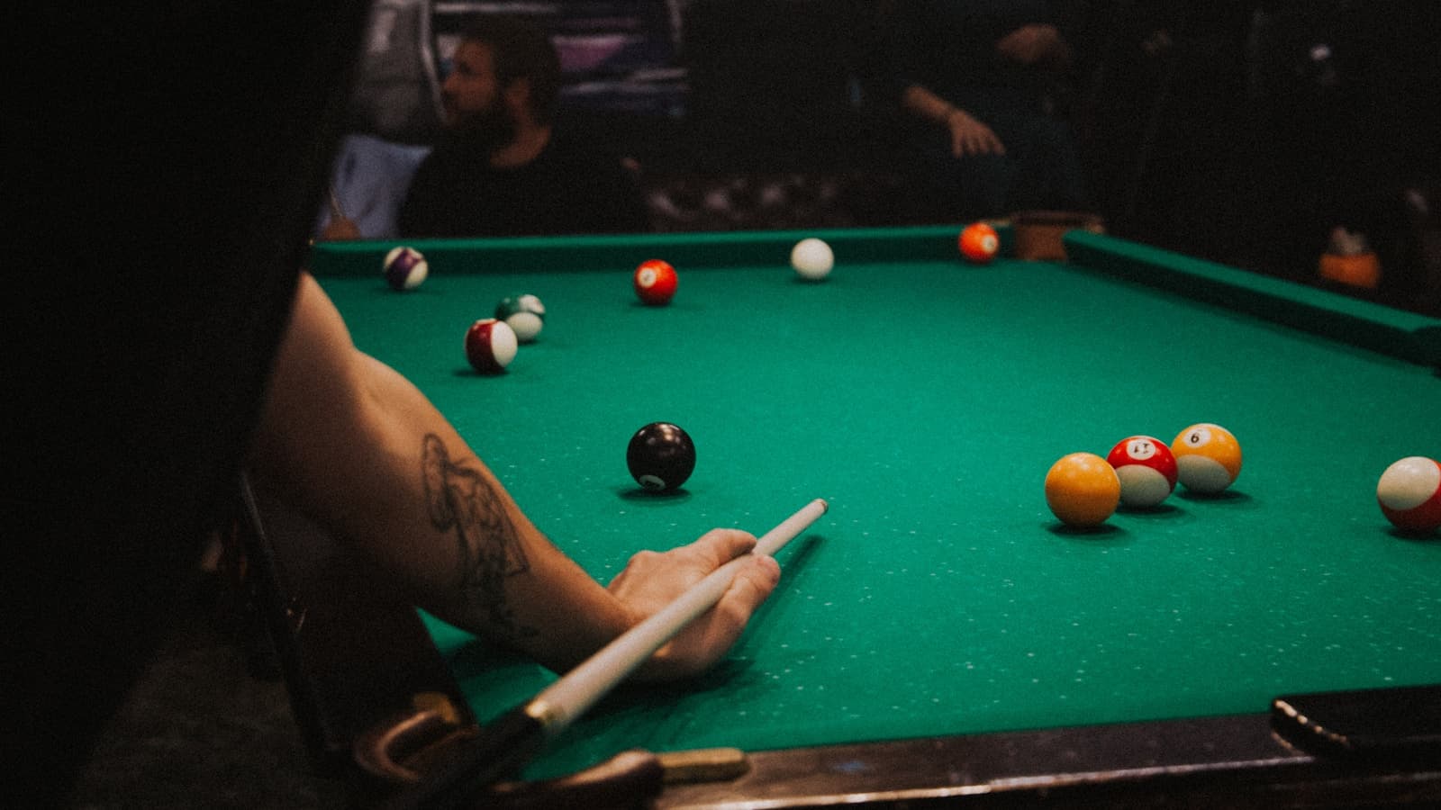 An arm and cue reaching over a green felt pool table with balls scattered mid-game.