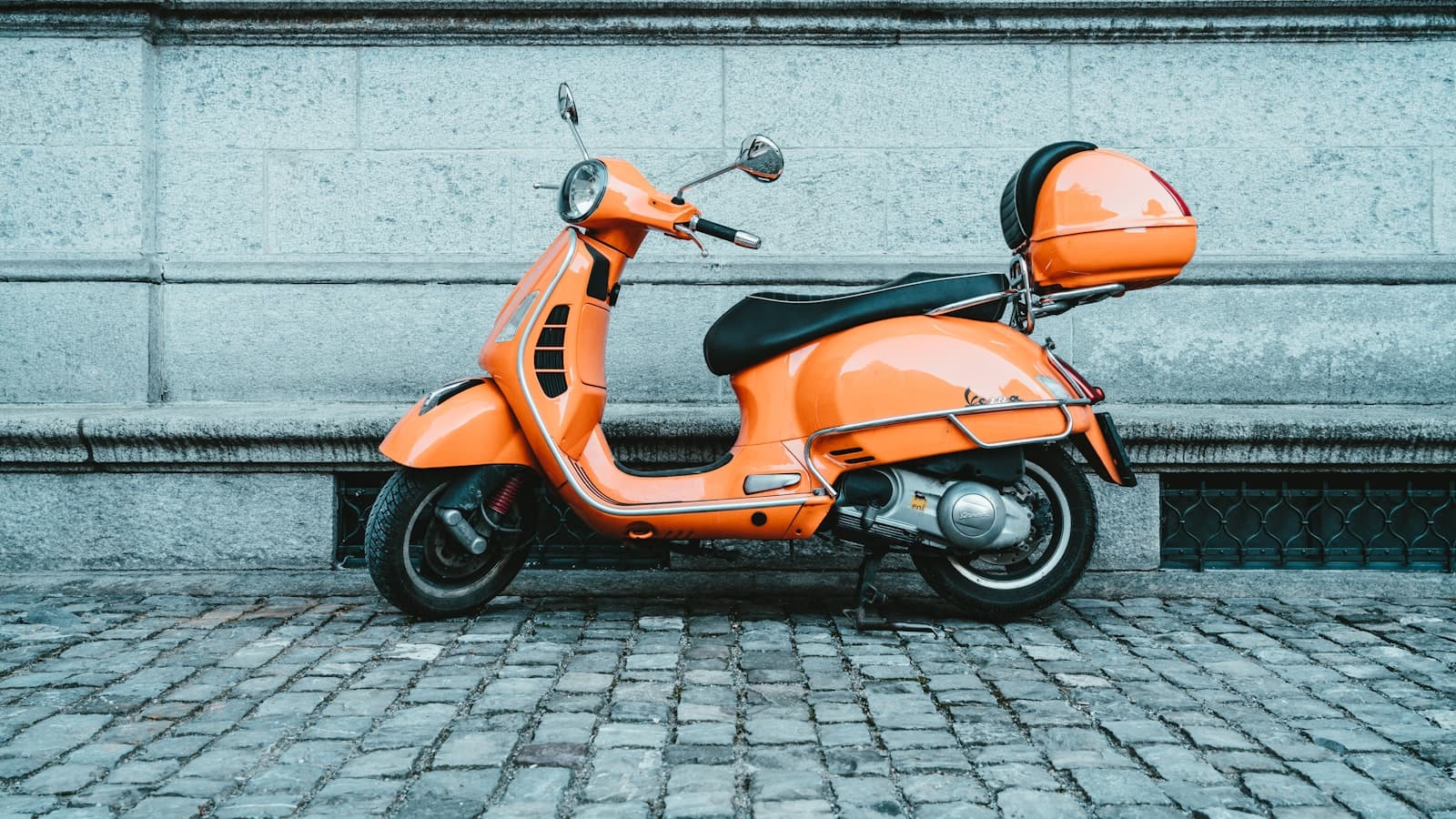 An orange motor scooter parked against a stone wall on a cobblestone street, no people, text, or brand markings visible.