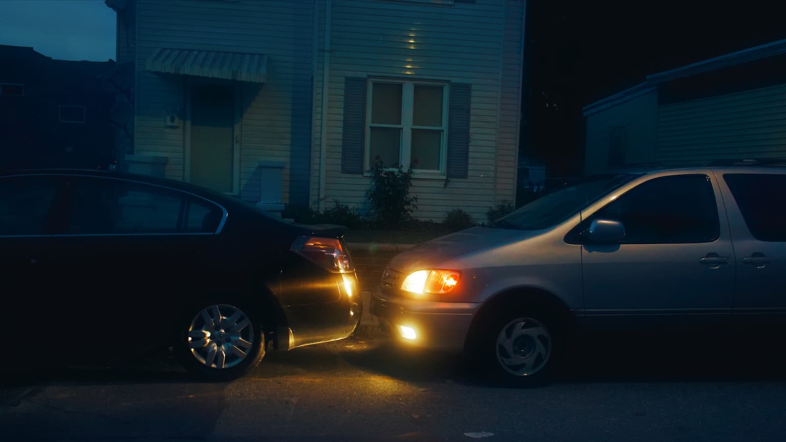 Two ordinary cars nose-to-nose in a residential driveway at dusk with headlights on, with no visible faces, plates, or brand logos.