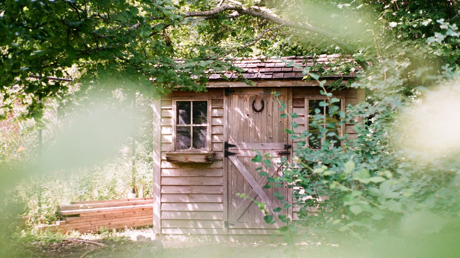 A charming wooden garden storage shed with windows and a horseshoe on the door, seen through soft-focus green foliage; no faces or text.