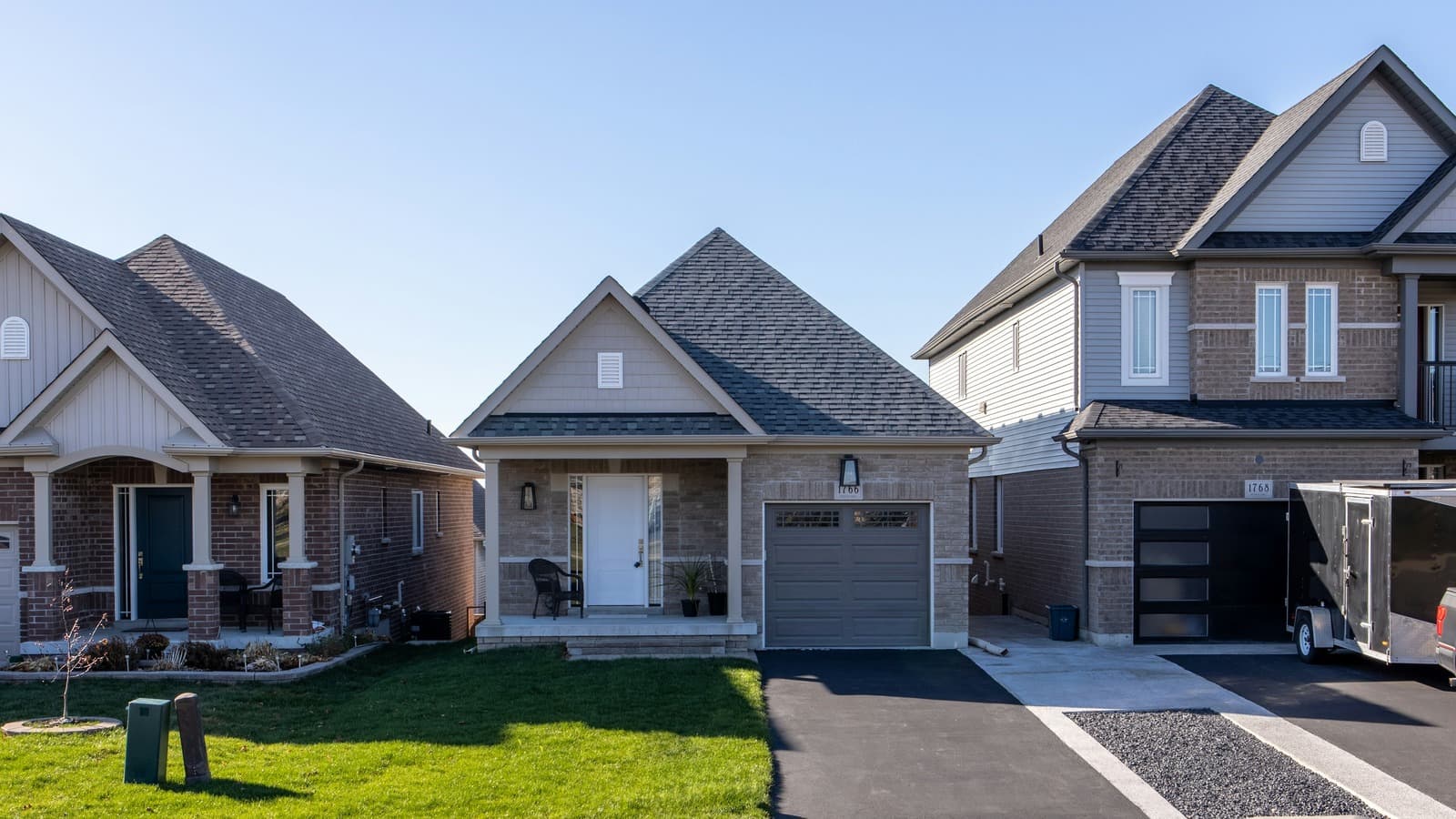 A small single-story brick starter home with a one-car garage, covered porch, and front lawn, flanked by larger neighboring houses on a suburban street.