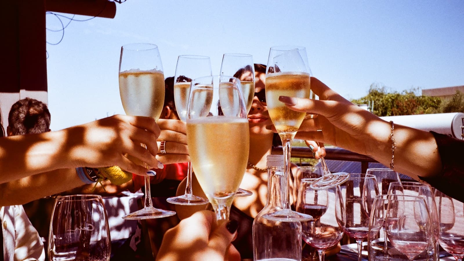A group of friends clinking champagne glasses together outdoors in the sun.