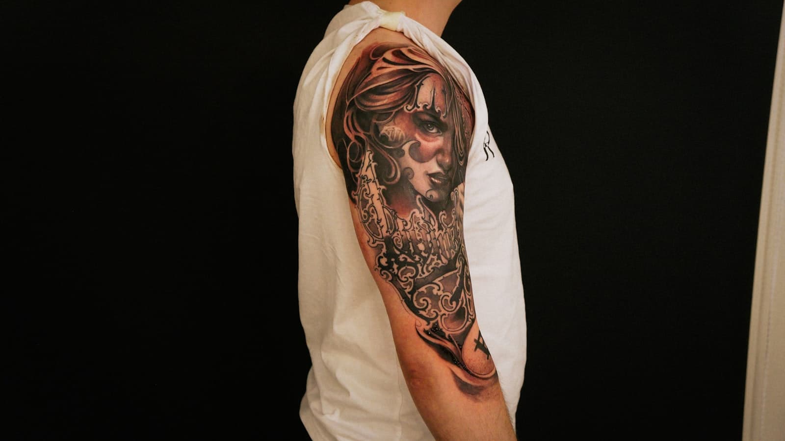 A close-up of a person's tattooed arm and shoulder sleeve against a black background, with no identifiable face visible.
