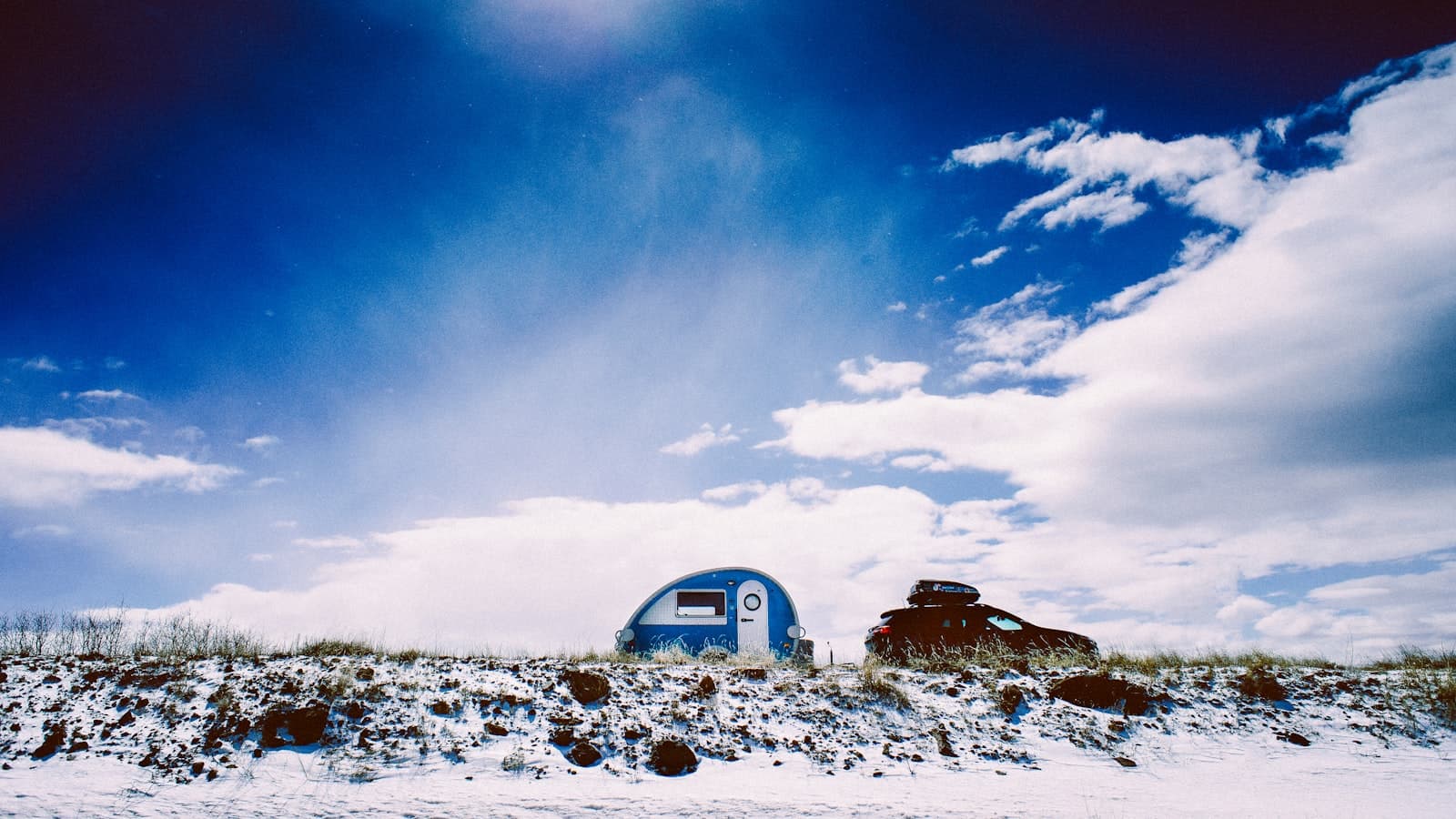 A small blue-and-white teardrop camper trailer parked next to a dark SUV on a snowy, grassy hilltop under a blue sky with clouds.