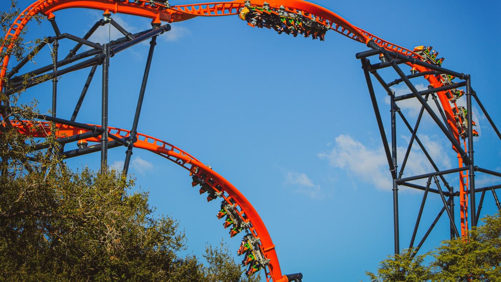 Vibrant orange roller coaster track against a blue sky with riders too small and distant to be identifiable, no text or logos.