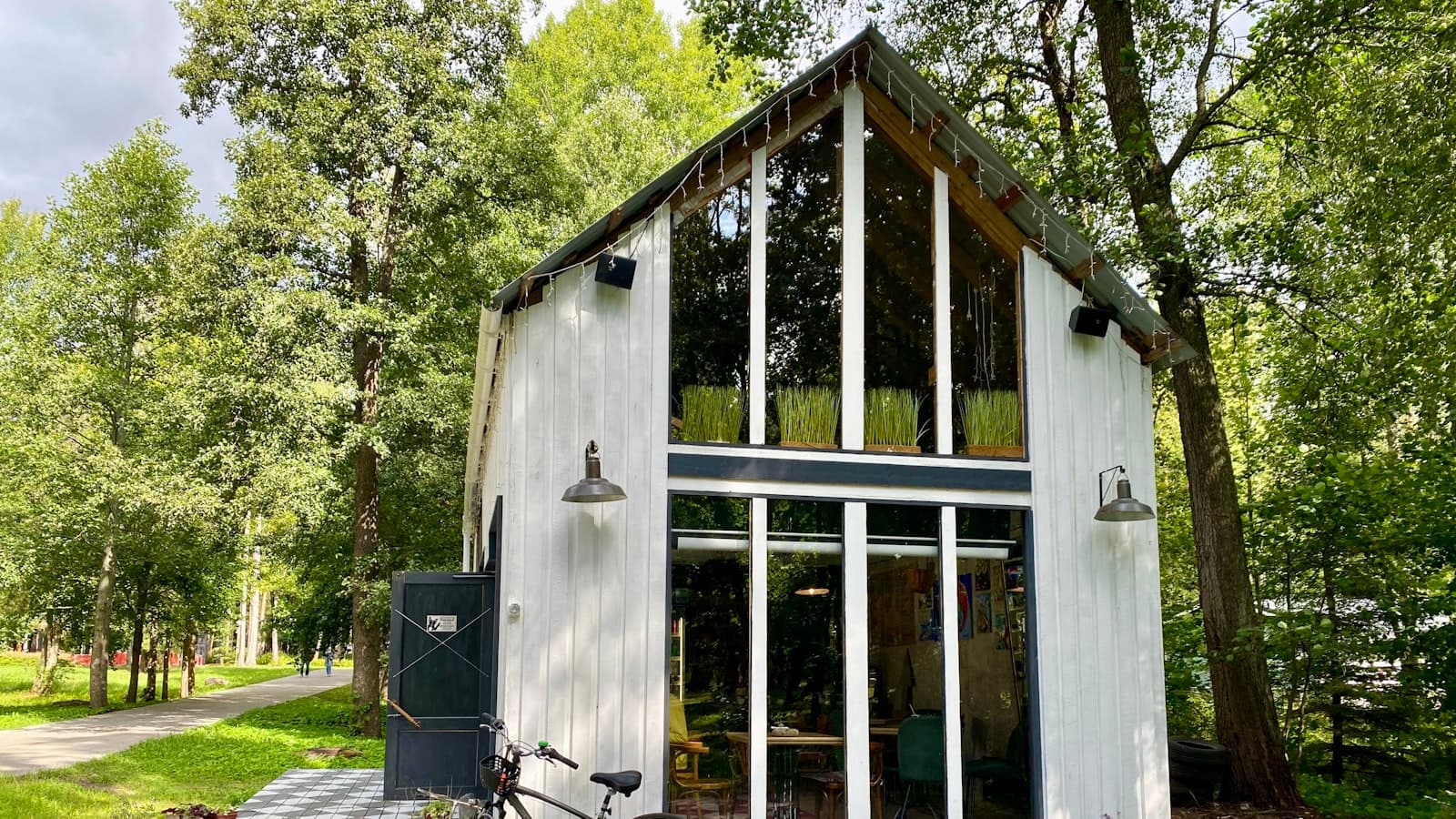 A modern white tiny home with a steep peaked roof and floor-to-ceiling windows surrounded by trees, with a bicycle parked out front.
