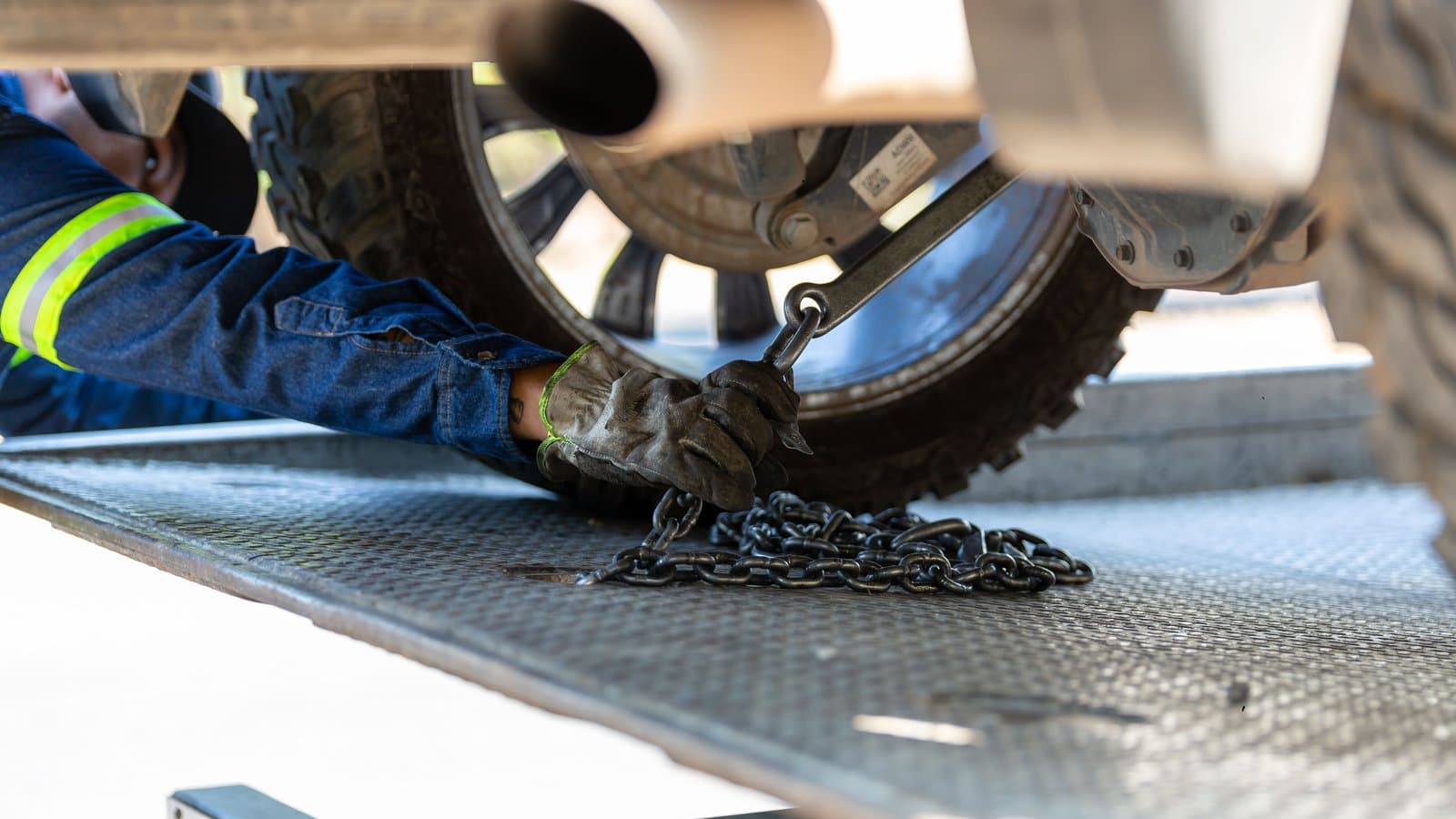 Close-up of a gloved hand attaching a tow chain and hook to the underside of a vehicle loaded on a flatbed tow truck.