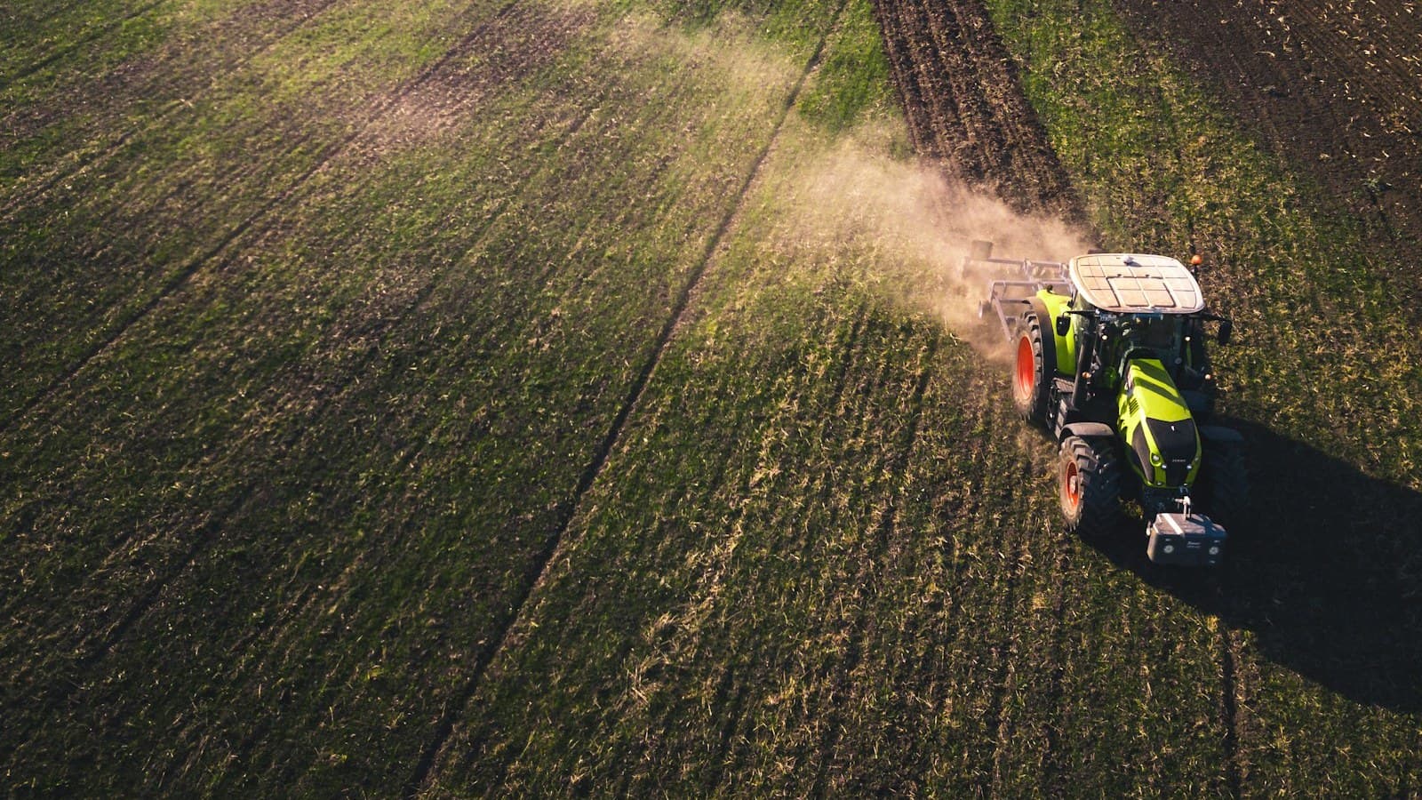An aerial shot of a green tractor plowing a field, kicking up dust, with no driver, readable branding, or plate visible.