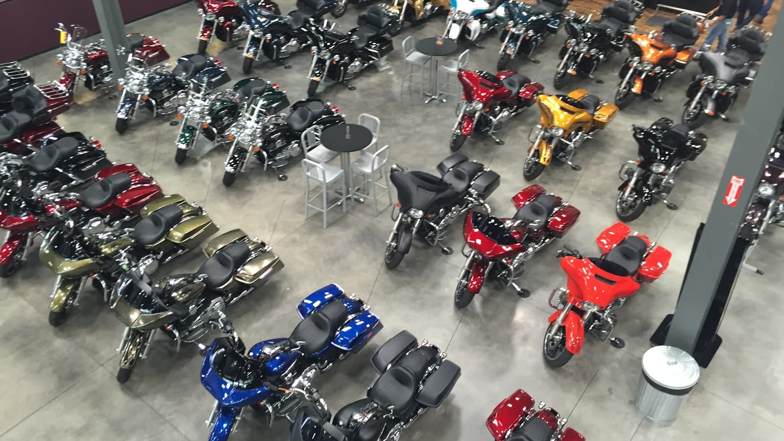 An overhead view of dozens of motorcycles arranged on a showroom floor, with no legible plates or logos and only distant, unidentifiable people in the far corner.