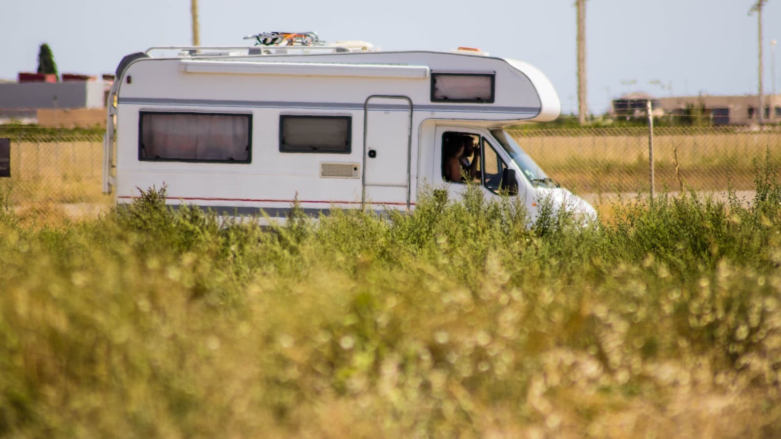 An older white Class C motorhome parked in a sunlit grassy field with a chain-link fence and low buildings in the background.