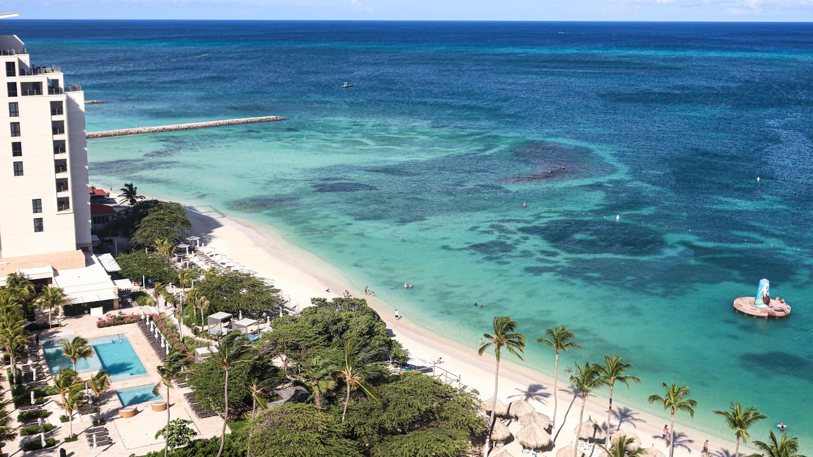 Aerial view of a resort beach with turquoise water and palm trees, people too distant to be identifiable and no readable signage.