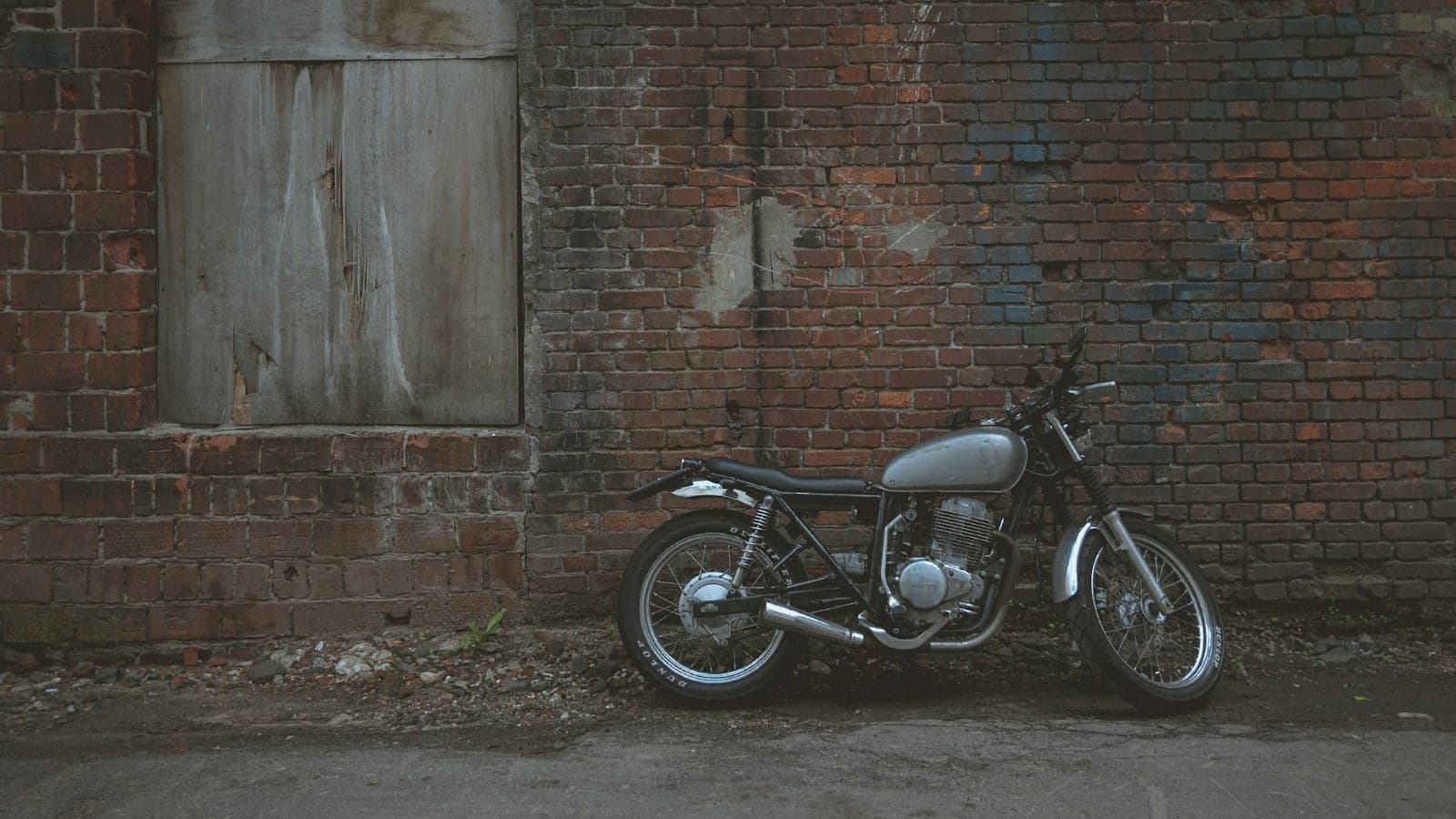 A weathered cafe-racer style motorcycle leaning against a worn brick wall, with no visible plate, rider, or legible brand logo.