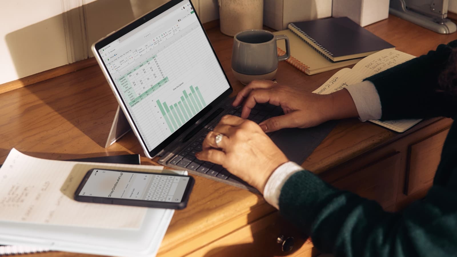 A person typing on a laptop showing a spreadsheet at a sunlit home-office desk.