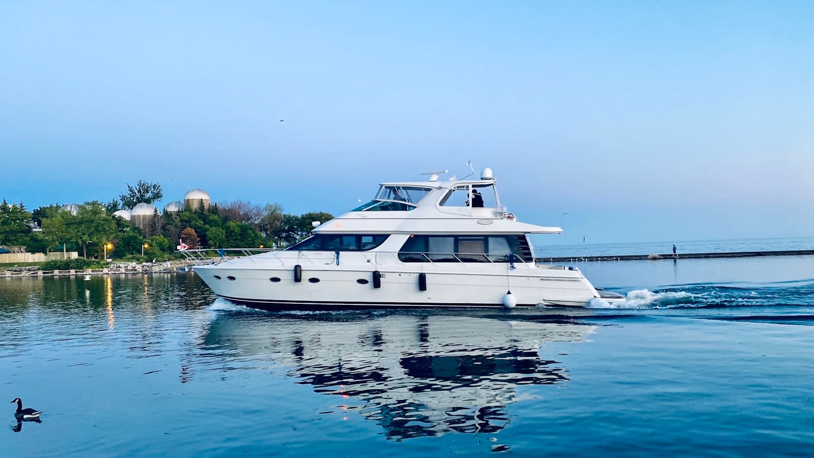 A mid-size white flybridge motor yacht cruising near a harbor at dusk, small unidentifiable silhouetted figures, no readable name or registration on the hull.