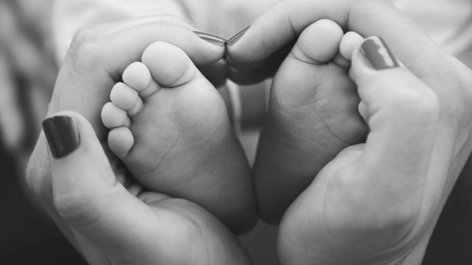 Black-and-white close-up of adult hands gently cupping a baby's feet into a heart shape, with a heavily blurred, unidentifiable figure in the background; no readable text.