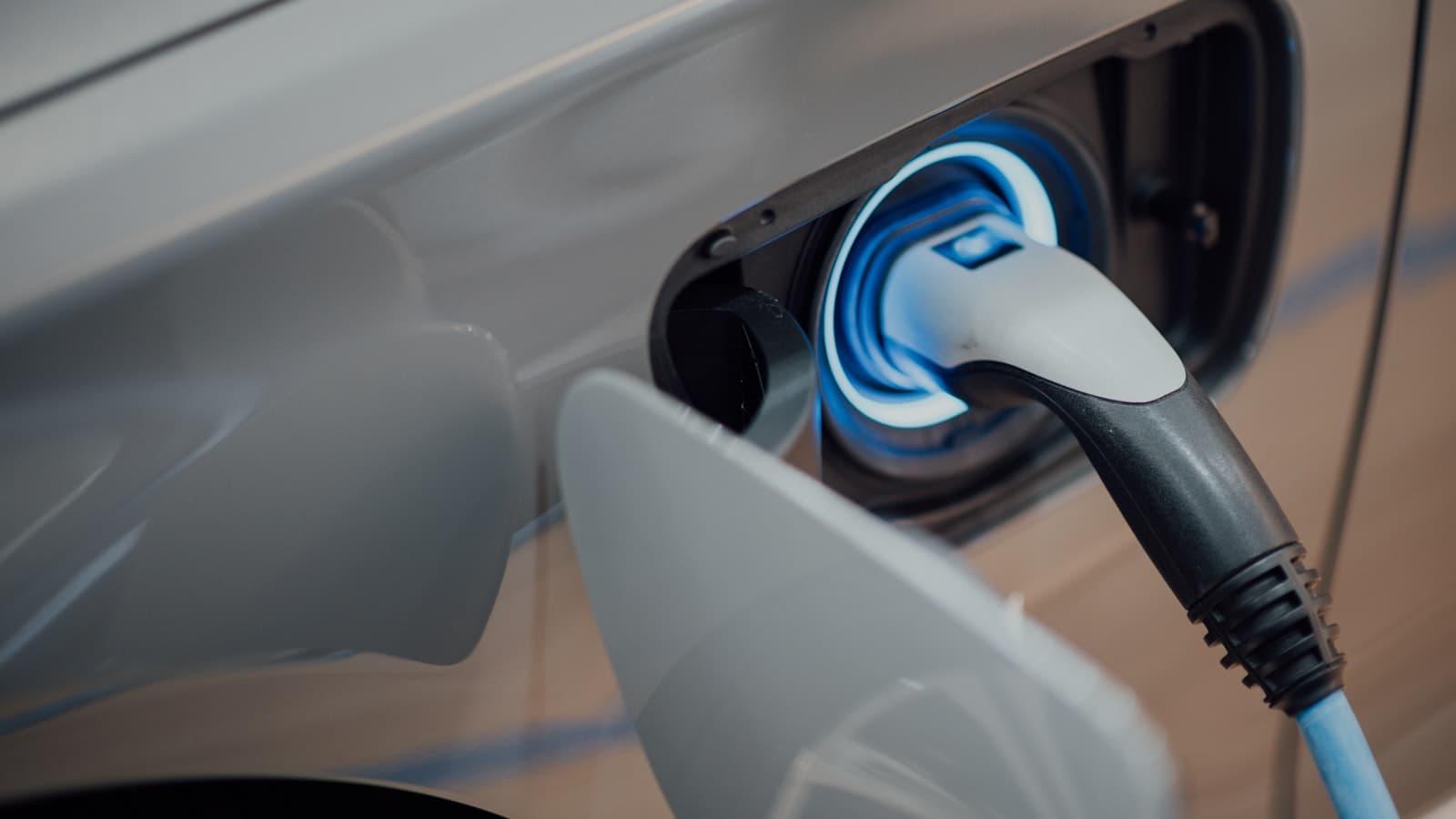 Close-up of a glowing blue charging cable plugged into a silver electric car's charge port, no faces, plates, or brand logos visible.